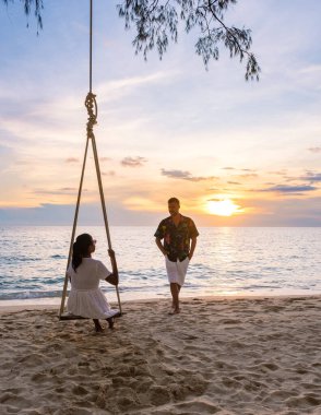 Kumsalda gün batımında salıncakta sallanan çift, Taylandlı kadınlar ve beyaz erkekler günbatımını, Tropikal Ada Koh Kood veya Koh Kut Tayland 'ı izliyor.. 