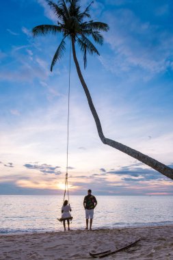 Koh Kood Tayland, plaj salıncaklı tropik bir sahil cenneti. Birkaç erkek ve kadın, güzel bir tropikal kumsal yaz tatilinde hindistan cevizi palmiyesinin altında salıncakta sallanıyor.