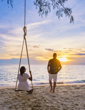 Kumsalda gün batımında salıncakta sallanan çift, Taylandlı kadınlar ve beyaz erkekler günbatımını, Tropikal Ada Koh Kood veya Koh Kut Tayland 'ı izliyor..