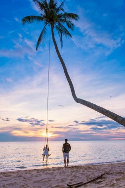 Kumsalda gün batımında salıncakta sallanan çift, Taylandlı kadınlar ve beyaz erkekler günbatımını, Tropikal Ada Koh Kood veya Koh Kut Tayland 'ı izliyor..