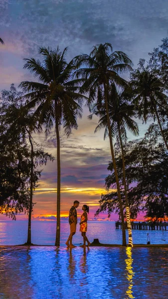 一对男女在热带游泳池边 棕榈树紧盯着夕阳 — 图库照片
