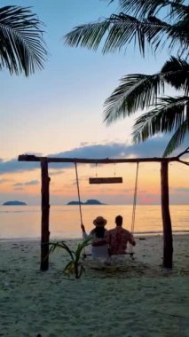 Tayland Koh Chang Adası 'ndaki bir salıncaktan gün batımını izleyen bir çift kadın ve erkek. Tropik bir plajda gün batımı. Asyalı Taylandlı kadınlar ve Avrupalı erkekler sahilde
