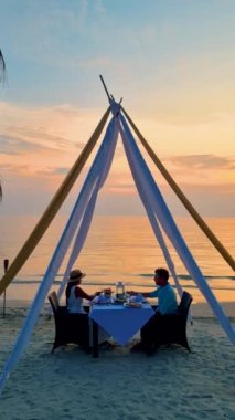 Koh Chang Tayland adasında gün batımında Tayland yemeğiyle sahilde romantik bir akşam yemeği. Birkaç beyaz erkek ve Asyalı Taylandlı kadın sahilde romantik bir akşam yemeği yiyorlar.