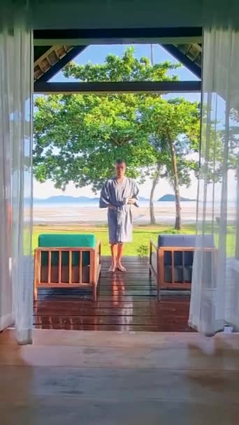 年轻人醒来后在热带海滩的一个海滨平房里喝咖啡 海滩上的豪华别墅里的年轻人 — 图库视频影像