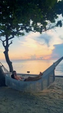 Hua Hin Tayland 'da tropik bir plajda güneşin doğuşunu izleyen bir çift kadın ve erkek. Huahin Tayland sahilindeki hamakta Asyalı kadınlar ve Avrupalı erkekler