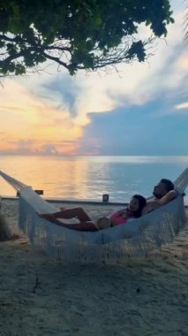 Hua Hin Tayland 'da tropik bir plajda güneşin doğuşunu izleyen bir çift kadın ve erkek. Huahin Tayland sahilindeki hamakta Asyalı kadınlar ve Avrupalı erkekler