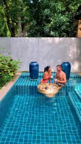一对夫妇在游泳池里吃早餐 亚洲女人和白人男人在游泳池里吃漂浮的早餐 — 图库视频影像