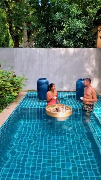 一对夫妇在游泳池里吃早餐 亚洲女人和白人男人在游泳池里吃漂浮的早餐 — 图库视频影像