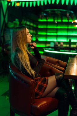 Uzun sarı saçlı, kısa etekli, siyah balıkçı yakalı, neon rengi açık yeşil bir restoranda, deri koltukta oturan genç ve çekici bir kadın.. 