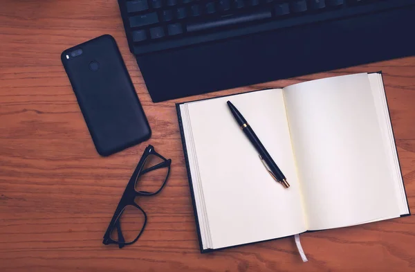 İş yeri. Tahta bir masanın üzerinde klavye, telefon, gözlük, defter ve kalem bulunur..