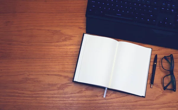 İş yeri. Tahta bir masanın üzerinde yer almaktadır: klavye, gözlük, defter ve kalem..