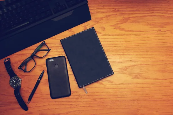 İş yeri. Tahta bir masanın üzerinde klavye, kol saati, gözlük, kalem, cep telefonu ve defter bulunmaktadır..