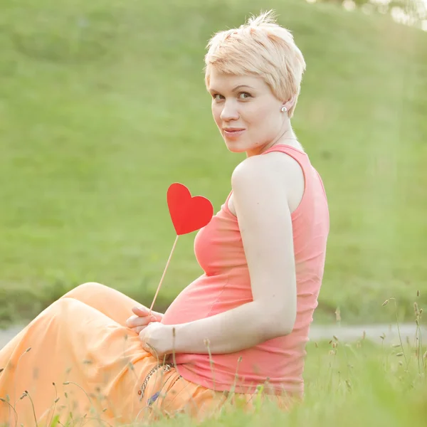 Pregnant woman enjoying summer park holding a paper red heart, o — Stock Photo, Image