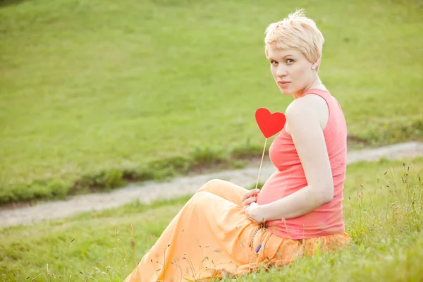 Gravid kvinna njuter av sommaren park håller ett papper rött hjärta, o — Stockfoto