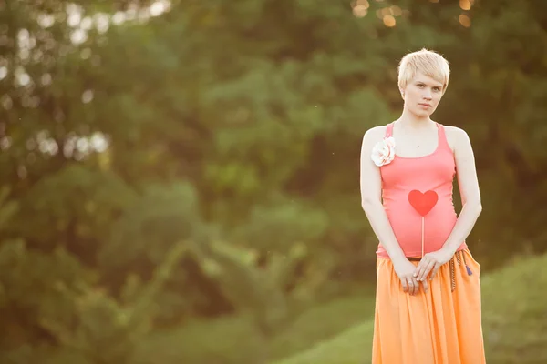 Mujer embarazada disfrutando del parque de verano sosteniendo un corazón rojo de papel, o —  Fotos de Stock