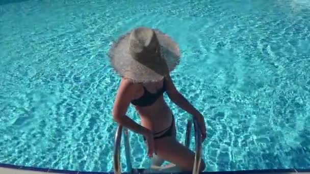 Młoda pewna siebie blondynka w czarnym stroju kąpielowym pobyt w basenie i patrząc na tle niebieskiej wody. — Wideo stockowe