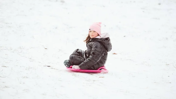 幸せな少女は、ソーサーそりで丘を滑り降りる。雪の上でスライダーに乗る女の子 — ストック写真