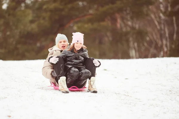 Ευτυχισμένη μητέρα με το κοριτσάκι να κάθεται στο έλκηθρο και να πέφτει με έλκηθρο στο χιόνι από το λόφο. Απολαμβάνω την άσπρη χειμωνιάτικη μέρα στο πάρκο. Περνώντας χρόνο μαζί το Σαββατοκύριακο. — Φωτογραφία Αρχείου