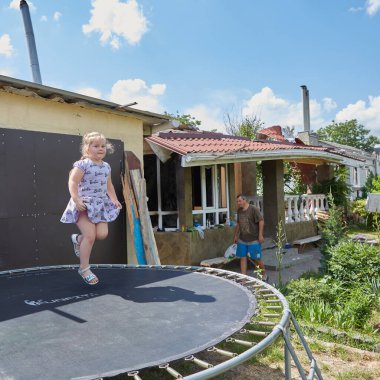 Chernihiv, Ukraine - June 26, 2022: Girl jumps on batute, against her destroyed house. Resident of the Chernihiv, whose house was completely destroyed by russian artillery. clipart