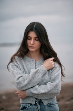 Bulutlu bir havada kumsalda tek başına duran uzun kahverengi saçlı kızın portresi. Deniz kenarı konseptinde bahar ya da sonbahar tatili. Kafkasyalı kadın portresi.