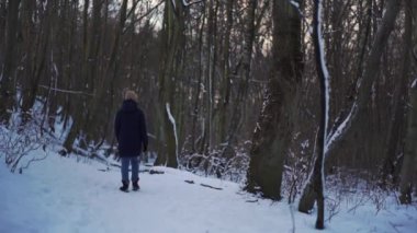 A person walking in winter forest. A shot of a woman back in the snowy forest. A girl walks slowly while wearing a cap and black jacket