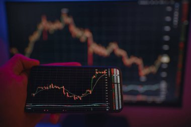 Arkaplanda bilgisayar ekranı olan şifreli döviz kurlarıyla akıllı telefon ekranı. Hisse senedi tüccarı, karlı fırsatları analiz ediyor. Bitcoin yatırıyor. Telefon ve bilgisayar kullanan kripto simsarı