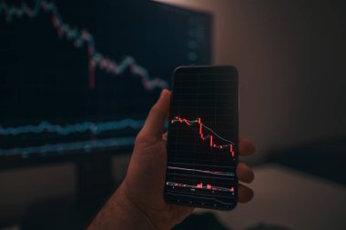 Arkaplanda bilgisayar ekranı olan şifreli döviz kurlarıyla akıllı telefon ekranı. Hisse senedi tüccarı, karlı fırsatları analiz ediyor. Bitcoin yatırıyor. Telefon ve bilgisayar kullanan kripto simsarı