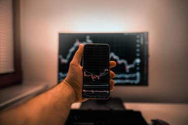 Arkaplanda bilgisayar ekranı olan şifreli döviz kurlarıyla akıllı telefon ekranı. Hisse senedi tüccarı, karlı fırsatları analiz ediyor. Bitcoin yatırıyor. Telefon ve bilgisayar kullanan kripto simsarı