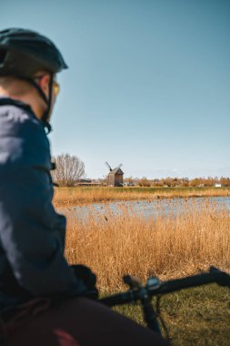 Nehir kıyısındaki klasik ahşap yel değirmeninin önünde miğferli siluet giyen bisikletçi. Aktif yaşam tarzı ve yaz boyunca seyahat kavramı. Kırsal alan manzarası