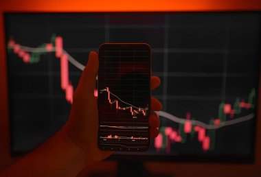 Arkaplanda bilgisayar ekranı olan şifreli döviz kurlarıyla akıllı telefon ekranı. Hisse senedi tüccarı, karlı fırsatları analiz ediyor. Bitcoin yatırıyor. Telefon ve bilgisayar kullanan kripto simsarı