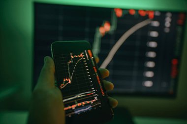 Arkaplanda bilgisayar ekranı olan şifreli döviz kurlarıyla akıllı telefon ekranı. Hisse senedi tüccarı, karlı fırsatları analiz ediyor. Bitcoin yatırıyor. Telefon ve bilgisayar kullanan kripto simsarı