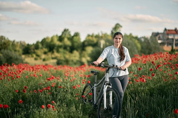 The girl stands in a white t-shirt with the bicycle in the red poppy field. Concept of active time in the countryside. Daily commuting with the bike during summer.