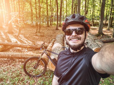 Ormanda kask ve güneş gözlüğü takan bir bisikletçinin selfie 'si. Dışarıda bisiklet sürerken gülümseyen bir adam. Kırsal bölgelerde bisiklet sürerken güvenlik kavramı