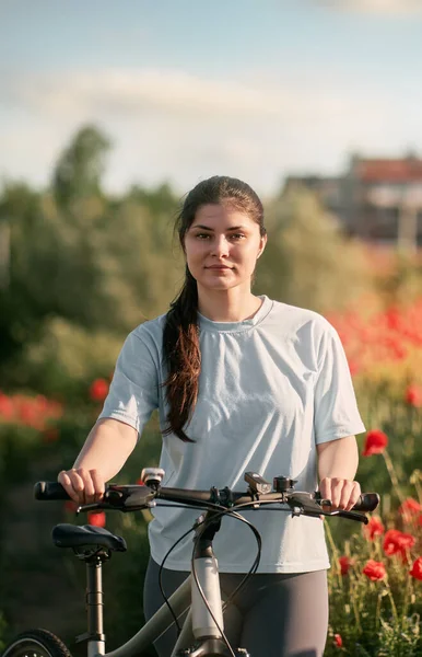 The girl stands in a white t-shirt with the bicycle in the red poppy field. Concept of active time in the countryside. Daily commuting with the bike during summer.