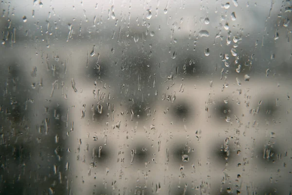 Raindrops on a window with an apartment house background. Concept of staying home while there is bad weather outside. Cozy indoor view at rainy weather