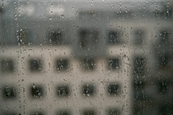 Rain droplets on the window glass. View from the window at the blurred street with buildings during bad weather.