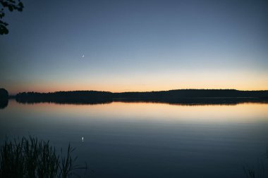 Gece mistik manzarası. Hilal ay, sisli gölün üzerinde diğer kıyıdaki ormanla ve durgun sulardaki yansımasıyla..