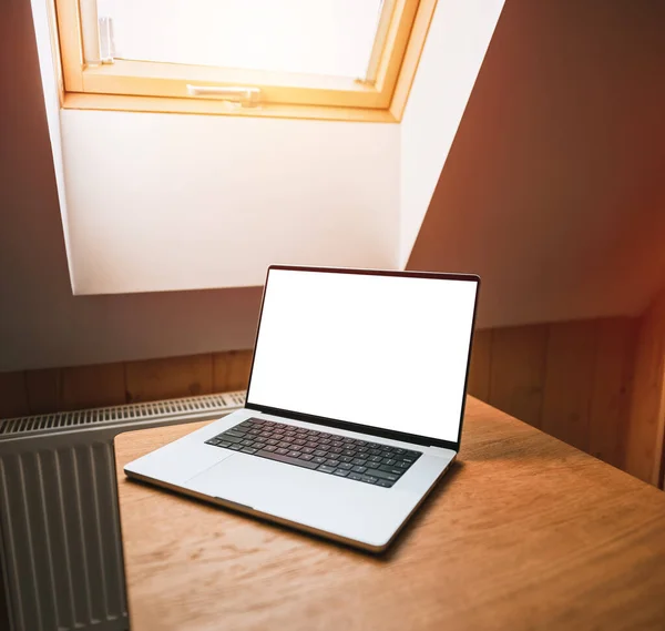 Opened modern professional laptop on a wooden table against opened roof window. Concept of working remotely in a cottage. Design mock-up with blank computer screen.