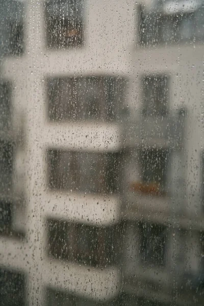 Raindrops on a window with an apartment house background. Concept of staying home while there is bad weather outside. Cozy indoor view at rainy weather