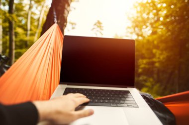 Design mock-up of a modern laptop computer with a blank white screen. Hiker is resting in a hammock in the woods. Using computer outdoors concept.