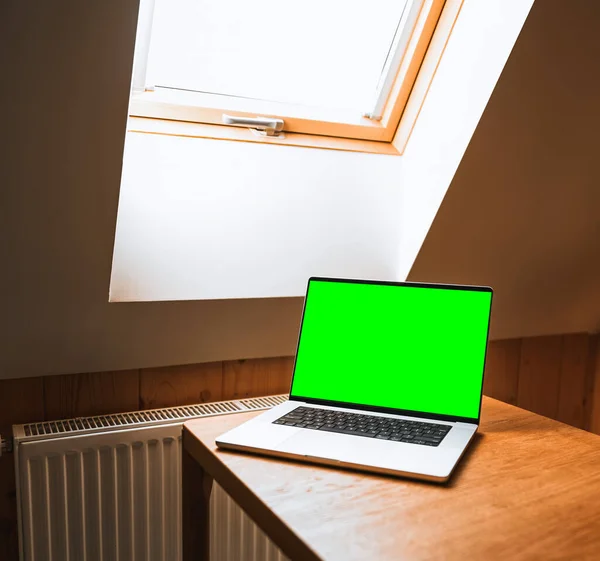 Opened modern professional laptop on a wooden table against opened roof window. Concept of working remotely in a cottage. Design mock-up with blank computer screen.