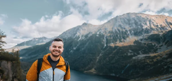 Man traveler smiles with beatufil mountain lake and forest in the background. Concept of outdoor adventures and hiking. Bearded young caucasian man wears backpack and sport outfit in the mountains.