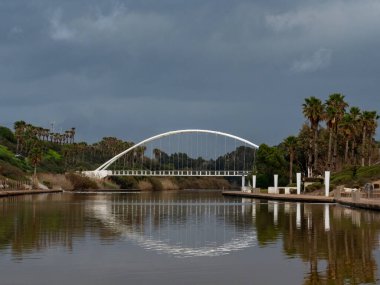Eğlence ve eğlence parkında nehrin üzerinde bir kavis köprüsü. Suda sağ taraftaki köprünün yansımasını görebilirsiniz, yürüyen bir alan. Gökyüzü bulutlu.