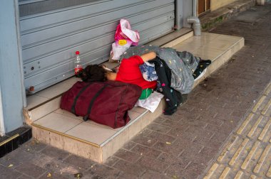 ATHENS, GREECE - MAY 14, 2022: A homeless man sleeps on a sidewalk in downtown Athens. Tourists pass by.