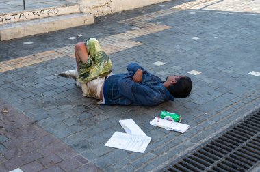 ATHENS, GREECE - MAY 14, 2022: A homeless man sleeps on a sidewalk in downtown Athens. Tourists pass by.