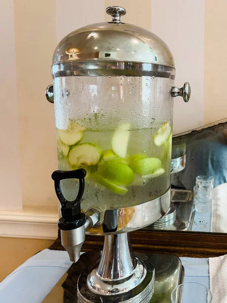 A glass transparent tank with a tap with cold drinking water with pieces of fruit and mint stands in the hotel lobby. Close-up.