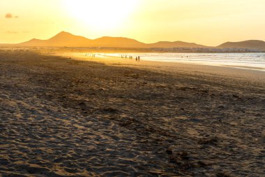İnsanların gün batımında yürüdüğü sahil manzarası Famara plajı, Lanzarote, Kanarya Adaları, İspanya.