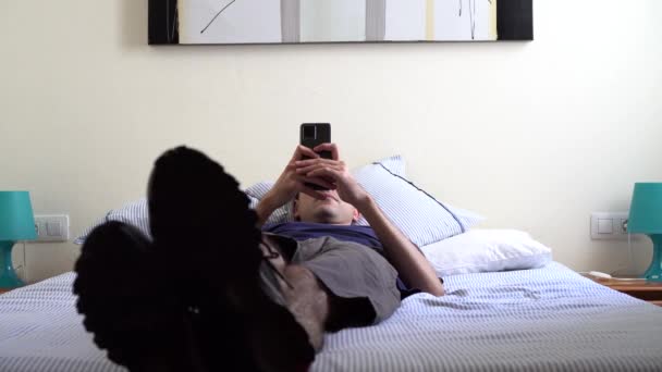 携帯電話を使って明るい部屋のベッドの上に横になっている若い男 — ストック動画
