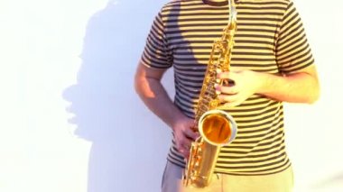 Genç adam açık havada beyaz bir duvarın önünde saksafon çalıyor..