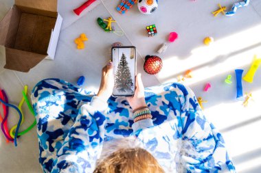 Çocuk oyuncaklarla oturuyor ve yılbaşı ağacı fotoğrafı ile akıllı telefon kullanıyor.